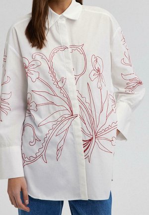 Chemise blanche boutonnée avec broderie florale rouge portée par une personne aux longs cheveux bruns, associée à un jean bleu.