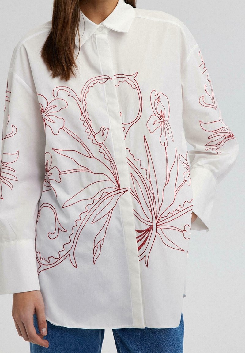 Chemise blanche boutonnée avec broderie florale rouge portée par une personne aux longs cheveux bruns, associée à un jean bleu.