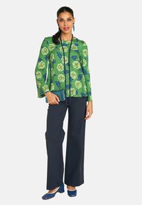 Frau in einem grünen, floral gemusterten Oberteil und weiten, marineblauen Hosen, die eine blaue Clutch hält. Sie hat lange Haare und trägt Ohrringe als Accessoire.
