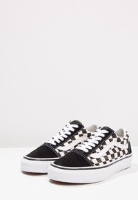 Baskets basses avec motif damier noir et blanc, lacets blancs et semelles épaisses en caoutchouc blanc sur fond uni.