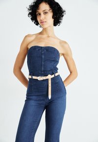 Jeans kombinezon v temno modri barvi brez naramnic, z oprijetim korzetom in hlačami visokega pasu, pritrjenimi z pasom iz svetlega usnja.