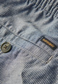 Tissu rayé bleu avec une finition texturée, présentant un bouton vert et une étiquette noire "SUPERDRY" cousue.