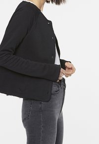 Black cropped jacket with a round neck, long sleeves, and raw hem. Styled with a white top and gray fitted jeans. Made from a soft fabric.