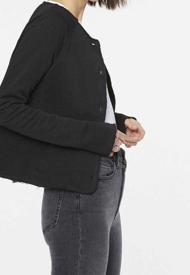 Black cropped jacket with a round neck, long sleeves, and raw hem. Styled with a white top and gray fitted jeans. Made from a soft fabric.