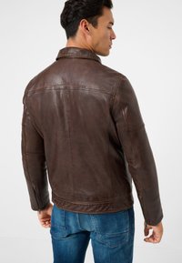 Bruine leren jas met een gestructureerde kraag, achternaad en ritsmanchetten, gecombineerd met blauwe denim jeans. Glad, textuurafwerking.