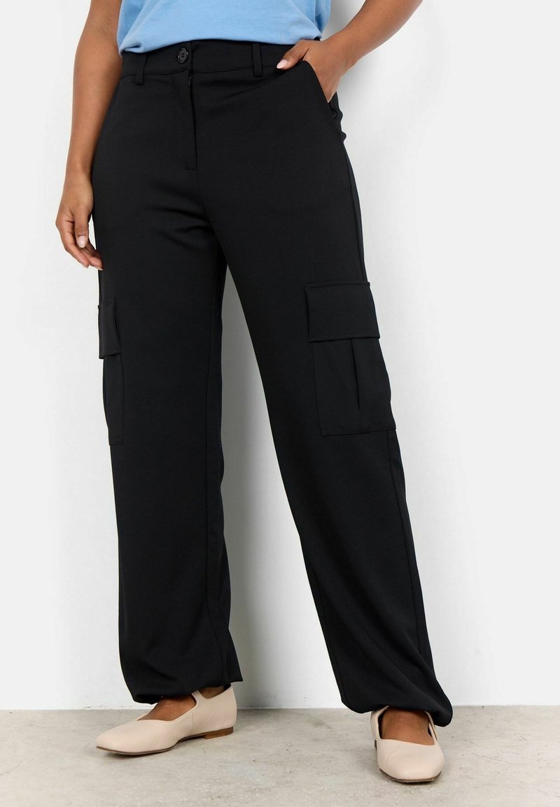 Personne portant un pantalon cargo noir avec de grandes poches latérales et des chaussures plates beiges, debout devant un mur blanc.
