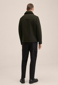 Giacca in pile verde scuro con colletto alto, abbinata a pantaloni cargo neri. Il design è strutturato, con cuciture e tasche visibili.