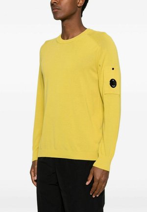 Uomo che indossa un maglione giallo a maniche lunghe con una toppa circolare nera sulla parte superiore della manica sinistra e pantaloni neri, in piedi su sfondo bianco.