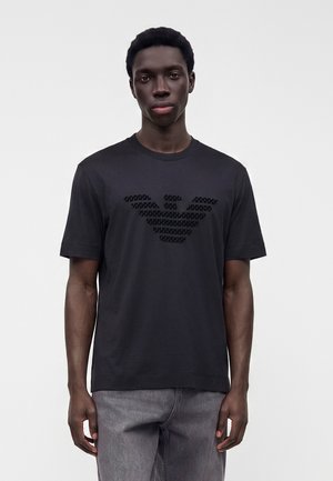 Jeune homme noir portant un T-shirt noir à manches courtes avec un logo d'aigle texturé et un jean gris, debout devant un fond clair uni.