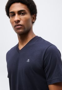 Νεαρός άνδρας με κοντά, σγουρά μαλλιά που φοράει μπλε ναυτικό T-shirt με V λαιμόκοψη και ένα μικρό λευκό λογότυπο «M OP» στο στήθος.