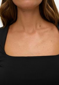 Zwarte top met vierkante halslijn van gladde, rekbare stof. Close-up toont de halslijn en sleutelbeen met minimale zichtbare details.