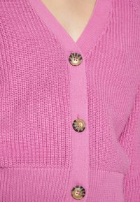 Różowy sweter na drutach z dekoltem w serek, pięcioma okrągłymi guzikami o kontrastowym wzorze oraz prążkowaną fakturą na krawędziach.