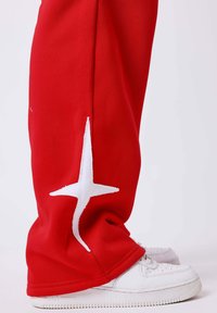Pantalon rouge avec un motif en forme d'étoile blanche près de la cheville, porté avec des baskets blanches sur un fond clair uni.