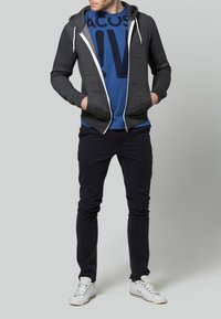 Grå zip-up hoodie med vita snören över en blå t-shirt med grafik. Mörka byxor och vita sneakers fulländar outfiten. En enkel grå bakgrund.