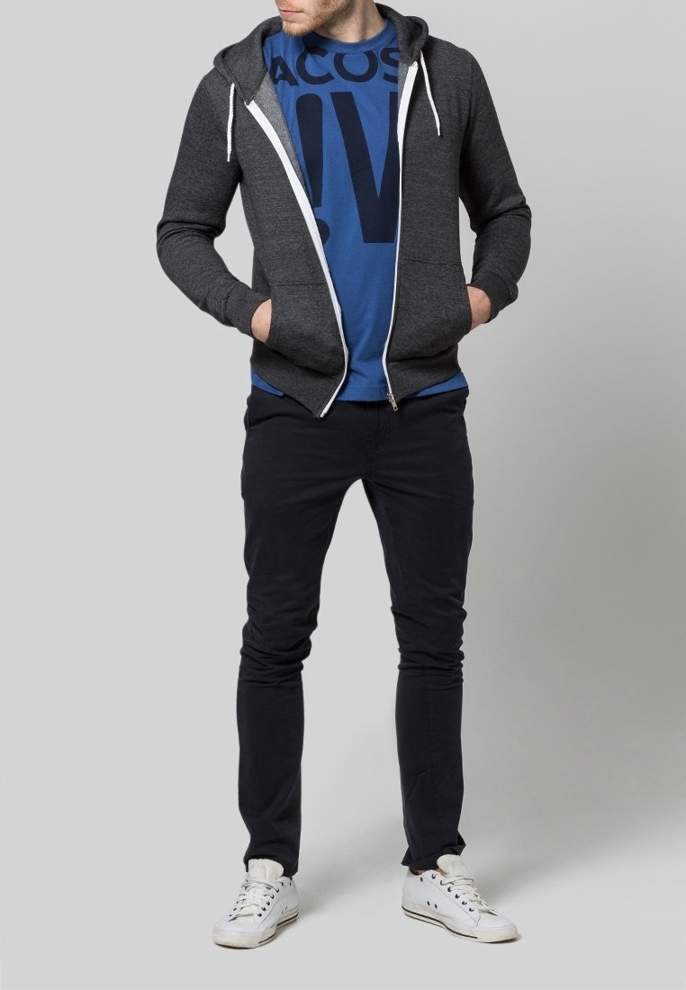 Grå zip-up hoodie med vita snören över en blå t-shirt med grafik. Mörka byxor och vita sneakers fulländar outfiten. En enkel grå bakgrund.