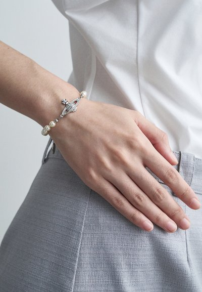 Mano descansando en la cadera, llevando una pulsera de encanto de plata con cuentas de perlas, combinada con una camisa blanca y pantalones grises claros texturizados.