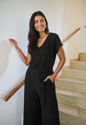 Kvinde med langt, mørkt hår iført en sort jumpsuit med bælte, stående ved en trappe, smilende og rørende ved et trægelænder.