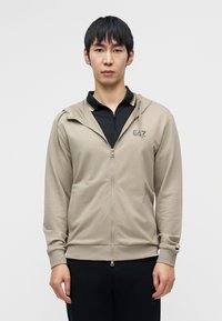 Μπεζ φούτερ με φερμουάρ και μπροστινές τσέπες, μαλακό ύφασμα, κουκούλα και διακριτικό λογότυπο. Φορεμένο πάνω από μαύρο πουκάμισο με γιακά και κίτρινα στοιχεία.