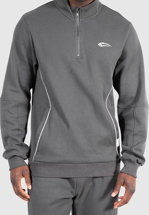 Mann trägt einen grauen Pullover mit Frontreißverschluss, weißen Paspeldetails und kleinem weißen Logo auf der Brust, kombiniert mit passenden grauen Hosen.