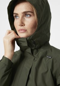 Donkergroene waterdichte jas met hoge kraag en verstelbare capuchon. Beschikt over een logo-tag aan de zijkant en een gladde, matte textuur.