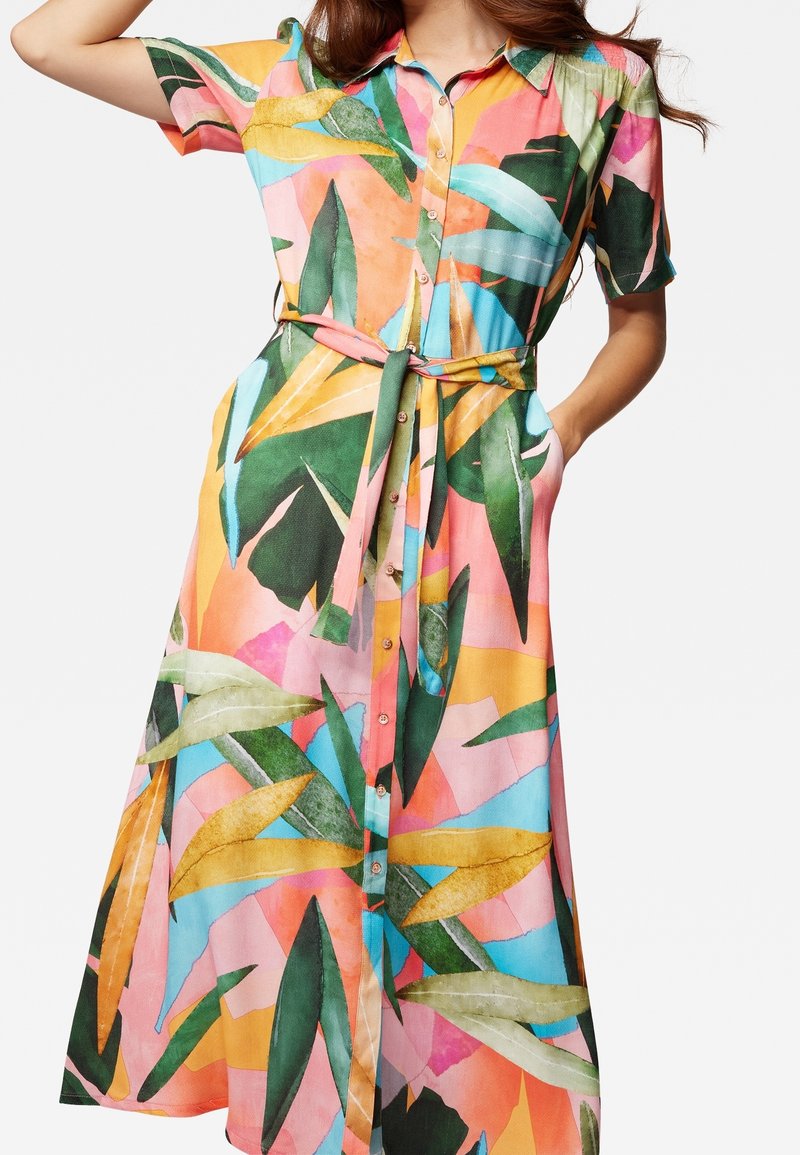 Femme portant une robe-chemise colorée à boutons avec un motif de feuilles tropicales et une ceinture nouée à la taille, la main reposant dans la poche.