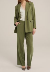 Olivově zelený přizpůsobený blazer s klopami a jedním knoflíkem, spárovaný s širokými kalhotami ve stejné barvě. Přes bílý top s krajkovými detaily.