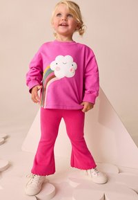 Rosa Sweatshirt mit einer Wolken- und Regenbogen-Grafik, kombiniert mit passenden gerippten Schlaghosen und weißen Sneakern.