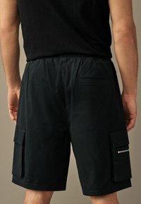 Bermuda cargo noir en tissu léger avec une taille élastique, des poches latérales et une poche arrière avec fermeture éclair.