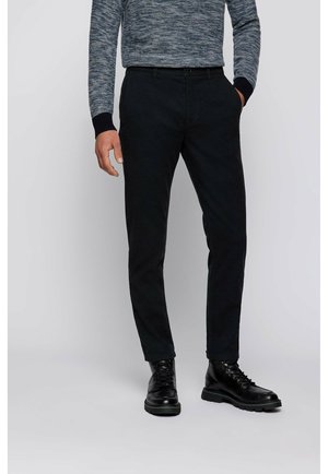 Mann trägt schwarze Slim-Fit-Hose, schwarze Leder-Schnürstiefel und einen blau-grauen Langarmpullover, steht mit einer Hand in der Hosentasche.