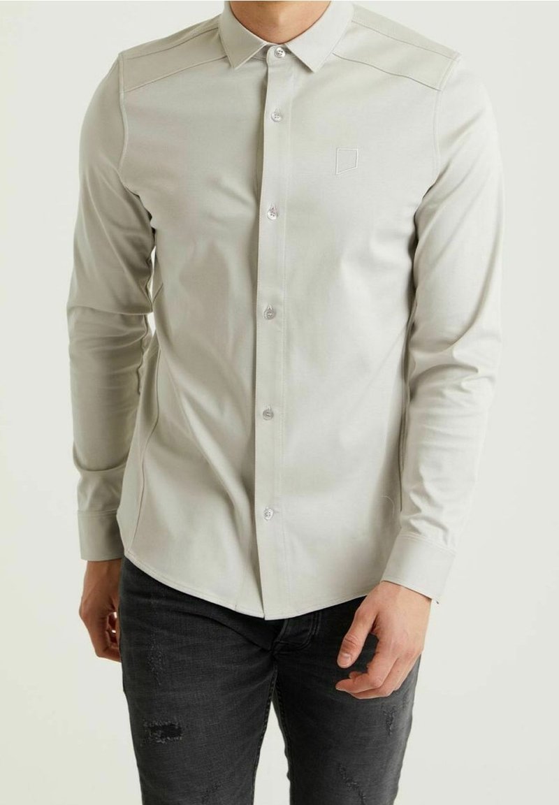 Homme portant une chemise ajustée gris clair à boutons et un jean noir déchiré, sur fond uni.