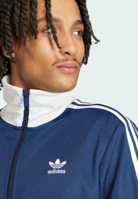 Dunkelblaue Zip-Jacke mit weißem Kragen und drei weißen Streifen auf den Schultern; verfügt über ein gesticktes Adidas-Logo auf der Brust.