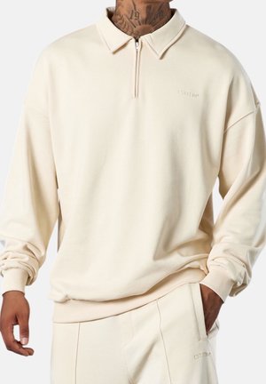 Mand iført beige langærmet kvart-lyn pullover med krave og matchende beige bukser, hånd i lommen, synlige nakkile tatoveringer.