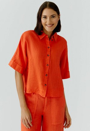 Femme aux cheveux châtain mi-longs souriante, portant une chemise ample boutonnée orange vif et un pantalon assorti, sur un fond clair.