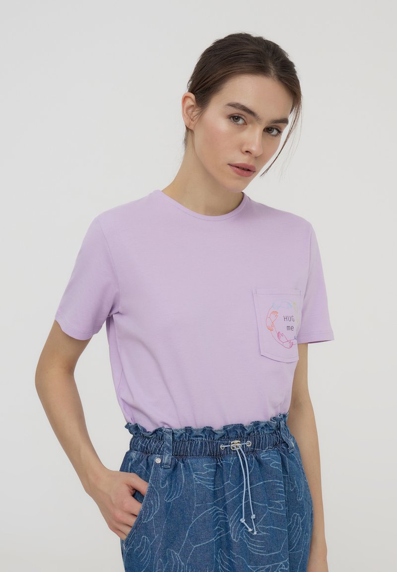 T-shirt in cotone lavanda con maniche corte, scollo rotondo e tasca con design "HUG me". Abbinato a pantaloni shorts in denim blu.