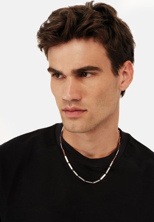Silvrfarvet metallinkekæde med rektangulære led, der har skiftevis polerede og matte overflader, båret over en sort T-shirt.