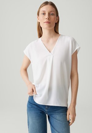 Frau trägt schlichte weiße kurzärmelige V-Ausschnitt-Bluse und blaue Jeans, steht vor neutralem Hintergrund mit einer Hand in der Tasche.