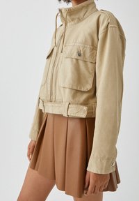 Beżowa krótka kurtka z wysokim kołnierzem, zapinana na zamek, z dwoma przednimi kieszeniami i epoletami na ramionach, noszona na brązowej spódnicy plisowanej.