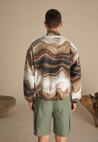 Jakke med abstrakt mønster i nuancer af brun og hvid, med elastisk kant og manchetter. Båret over grønne cargobukser, der viser letvægtsstof.