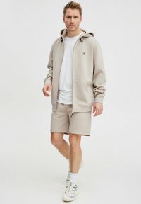 Haut de sweat à capuche zippé beige avec une capuche à cordon, associé à un short assorti. Le tissu est lisse avec un logo simple sur la poitrine.