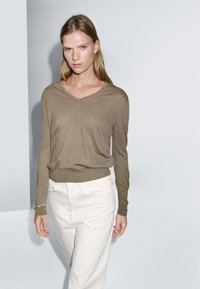 Olivegrüner V-Ausschnitt-Pullover aus weichem Material, mit langen Ärmeln und schmalem Saum, kombiniert mit weißen geraden Hosen.
