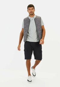 Grijze gewatteerde vest over een grijze polo. Marineblauwe cargoshorts, witte sneakers met marineblauwe accenten. Model loopt tegen een witte achtergrond.