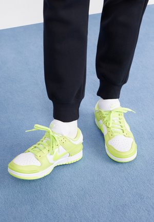Person iført sorte bukser, hvide sokker og hvide sneakers med neon grønne accenter, stående på en lyseblå tæppe.