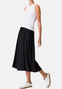 Jupe midi noire à taille à cordon, associée à un haut blanc sans manches. Le mannequin porte des baskets blanches avec des accents beige.