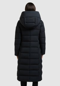 Kvinde iført en lang sort polstret dynejakke med stor hætte, set bagfra mod en ensartet lys baggrund.