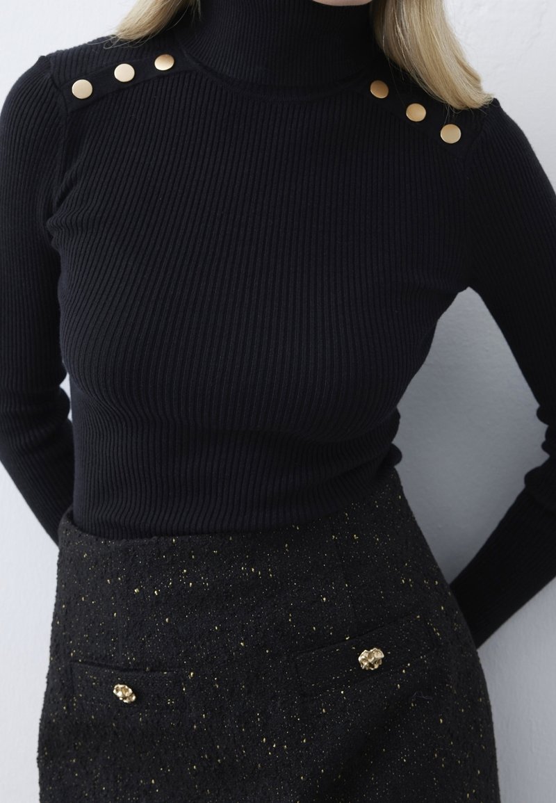 Schwarzer gerippter Rollkragenpullover mit drei goldenen dekorativen Knöpfen an der Schulter; kombiniert mit einem schwarzen Rock mit goldenen Sprenkeln und Akzenten.
