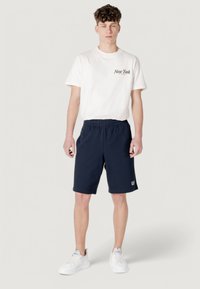 T-shirt en coton blanc avec le texte "New York", associé à un short bleu marine, doté d'une taille élastique et d'un détail de logo. Porté avec des baskets blanches.