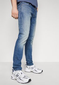 Férfi világoskék, koptatott slim-fit farmernadrágot és fehér New Balance sportcipőt visel, egy egyszínű világos háttér előtt.