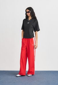 Γυναίκα που φορά μαύρο κοντομάνικο μπλουζάκι Puma, κόκκινο παντελόνι Puma φαρδύ με άσπρες ρίγες, ασημένια παπούτσια και σκούρα γυαλιά ηλίου, που στέκεται σε μπλε δάπεδο.