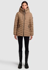 Tannfarbene Pufferjacke mit Kapuze, vertikaler Steppstruktur, Frontreißverschluss und zwei Seitentaschen. Kombiniert mit schwarzen Leggings und kniehohen Stiefeln.