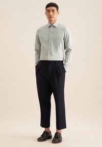 Camicia button-up verde chiaro con colletto a punta, abbinata a pantaloni plisati blu navy scuri e scarpe nere stringate, in piedi su uno sfondo neutro.